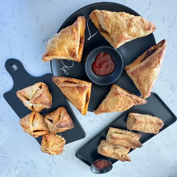 Assortment of golden-brown savory pastries, some triangular, some square, with small bowls of dipping sauce.
