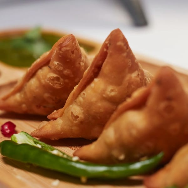 Three golden-brown samosas on a wooden board, garnished with green chili and a red berry.