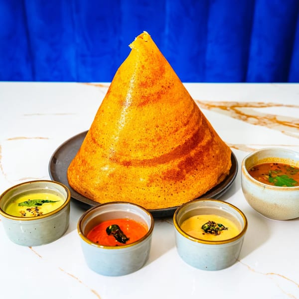 A golden-brown cone dosa stands tall on a plate, surrounded by four small bowls of colorful chutneys and sambar.