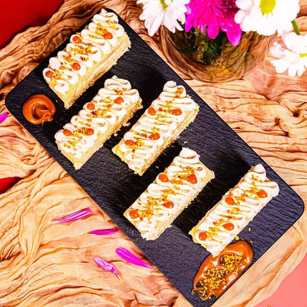 Five rectangular pastries with white frosting, caramel, and nuts on a black tray with flowers.