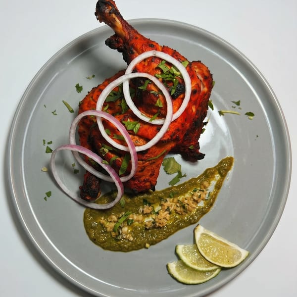 Tandoori chicken leg quarter with onion rings, green chutney, and lime wedges on a grey plate.