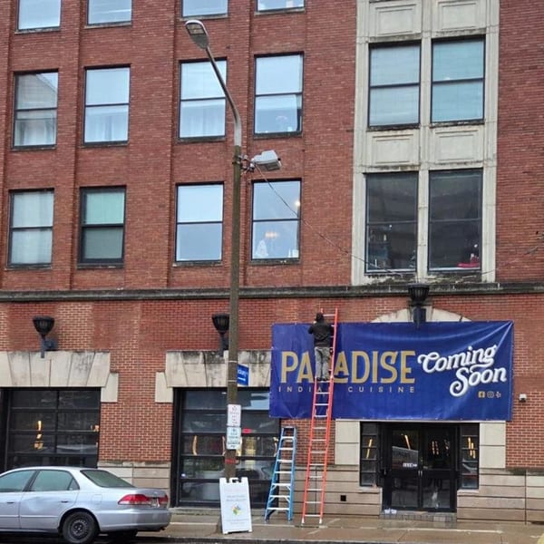 A person on a ladder installing a "Paradise Indian Cuisine Coming Soon" banner on a brick building.