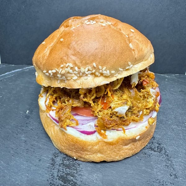 A pulled chicken sandwich with red onion, tomato, and white sauce on a sesame seed bun.