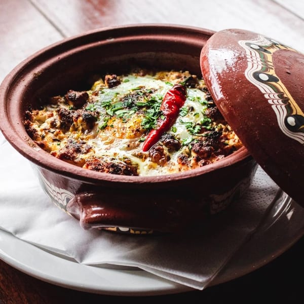 A clay pot filled with a baked dish, topped with herbs and a red chili pepper.